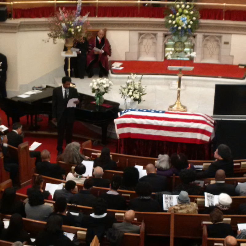 Haile Payne reads the obituary of his father, Les Payne, as the journalist’s casket sits wrapped in the American flag at Abyssinian Baptist Church in Harlem. The Rev. Dr. Calvin Butts III is at top. Former Newsday colleague Dele Olojede, a native of Nigeria, called Payne “first and foremost an American . . . He had a tremendous love for the country.”